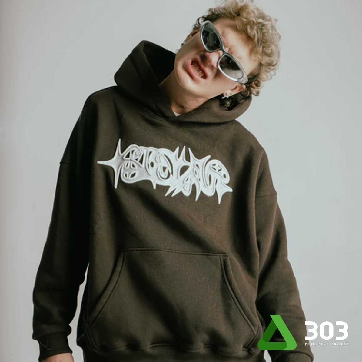 Osoba w brązowej bluzie z kapturem i białym nadrukiem, pozująca na jasnym tle. Nietypowe okulary i ekspresja twarzy dodają charakteru. Widoczne logo producenta odzieży.