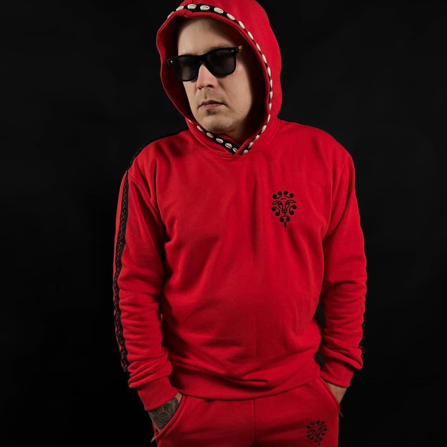 Mężczyzna w czerwonym dresie z kapturem, ozdobionym muszelkami, i czarnych okularach na tle czarnego tła. Na dresie widoczny czarny nadruk.