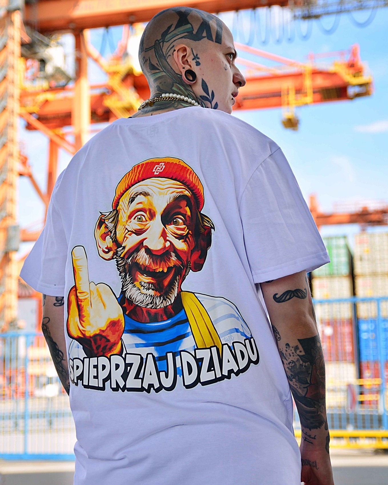 Mężczyzna z tatuażami prezentuje białą koszulkę z nadrukiem karykatury starszego mężczyzny w czapce i napisem. W tle dźwig portowy i kontenery.