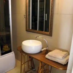 Biała, okrągła umywalka nablatowa na przerobionej, złotej podstawie starej maszyny do szycia, z chromowaną baterią i stosem beżowych ręczników w łazience z lustrem w ozdobnej ramie.