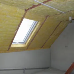 Żółta wełna mineralna zamocowana między krokwiami poddasza z oknem dachowym, zabezpieczona sznurkiem, widok na fragment ściany i drabiny.