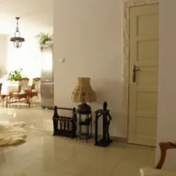 Jasne, błyszczące płytki podłogowe w przestronnym wnętrzu, z widoczną częścią stołu w pierwszym planie, lampą ze zdobionym abażurem, drewnianym stojakiem na gazety i figurką w tle, oraz drzwiami...