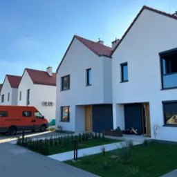 Nowoczesne szeregowe domy z czerwonymi dachami i pomarańczowym samochodem dostawczym zaparkowanym przed jednym z nich, widoczne trawniki i chodniki z kostki brukowej.