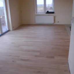 Jasna, nowo ułożona podłoga z desek w przestronnym pokoju z dużymi oknami balkonowymi i grzejnikiem pod oknem. Świeżo pomalowane ściany w odcieniach beżu.