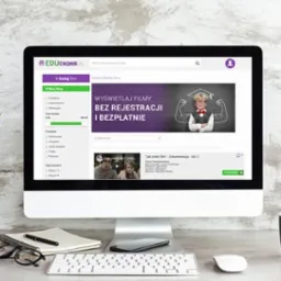 Ekran komputera iMac wyświetlający stronę internetową Educhomik.pl z bazą filmów edukacyjnych i scenariuszy lekcji, obok wazon z zieloną rośliną, notatnik, długopis, okulary i czarny kubek...