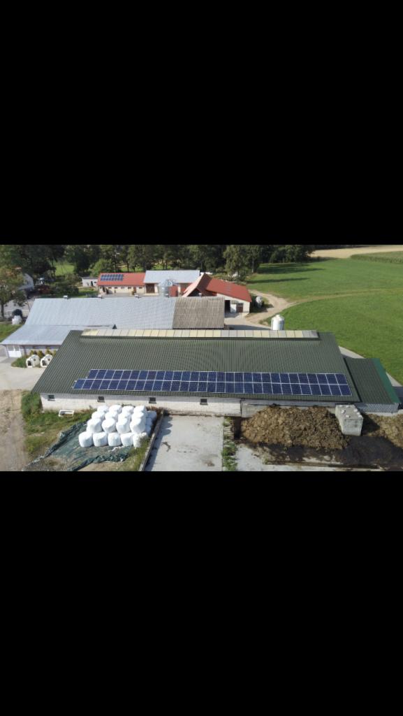 Widok z góry na budynek gospodarczy z panelami fotowoltaicznymi na dachu, obok stogi siana w białych osłonach, w tle zabudowania wiejskie i pole.
