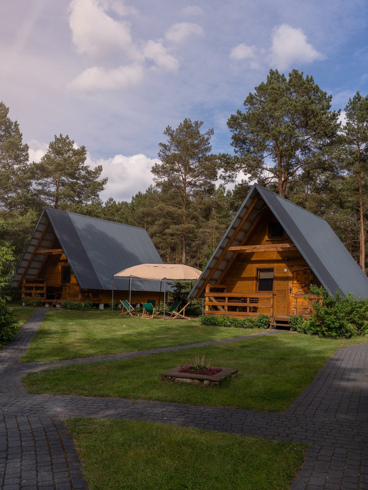 Dwa drewniane domki letniskowe z szarymi dachami w otoczeniu zieleni i drzew. Na trawniku parasol i leżaki. Ścieżka z kostki brukowej prowadzi do domków.