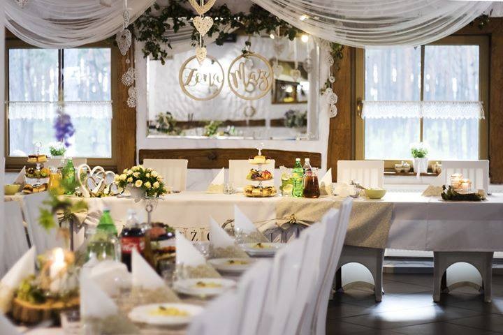 Uroczysty stół weselny z dekoracjami: kwiaty, napoje, talerze. Nad stołem wiszą ozdobne napisy 'Żona' i 'Mąż'. Okno w tle z białymi firankami.