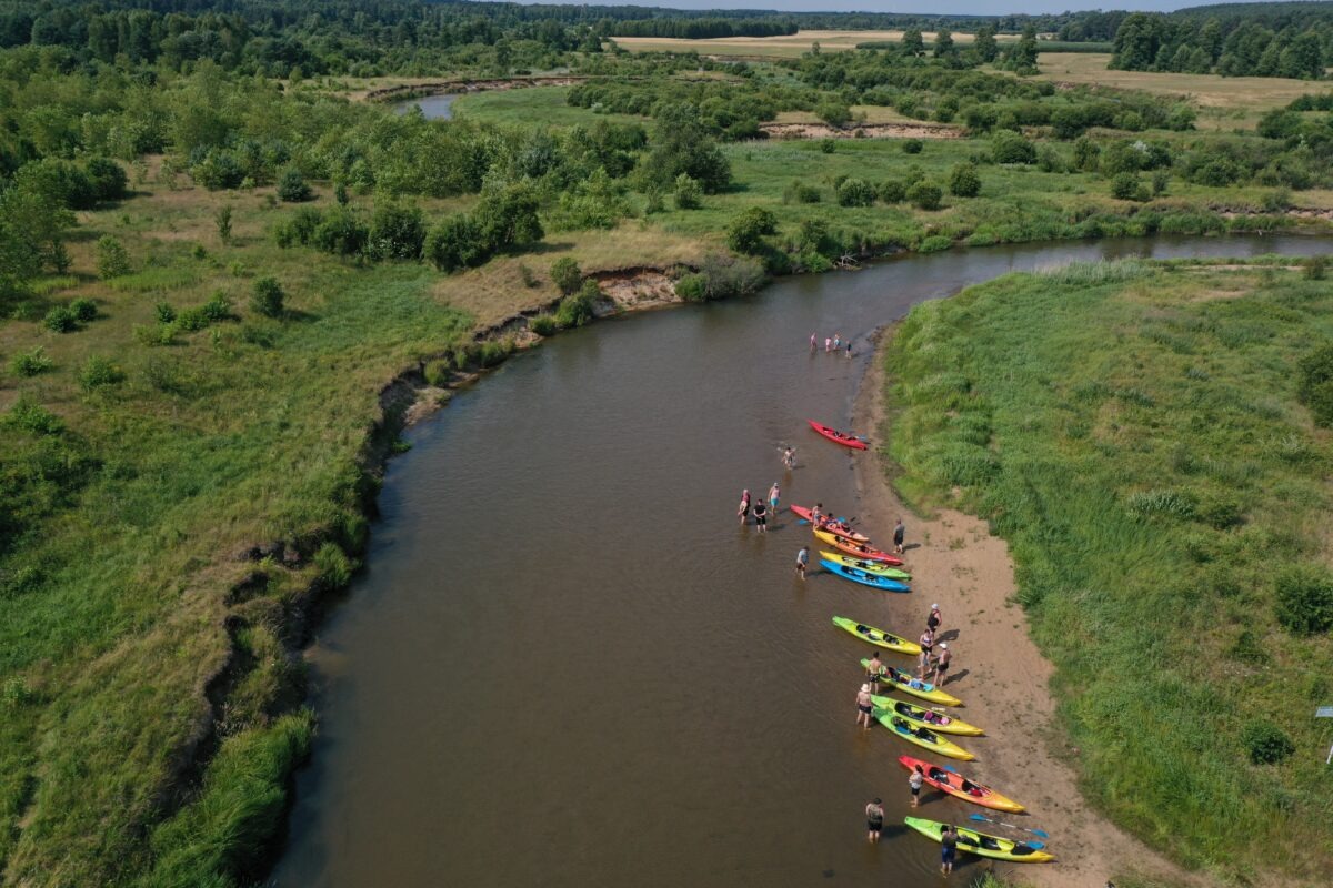 Widok z góry na rzekę z kajakami na brzegu i grupą ludzi w wodzie. Brzegi porośnięte zielenią, w tle las. Słoneczny dzień, rekreacja na świeżym powietrzu.