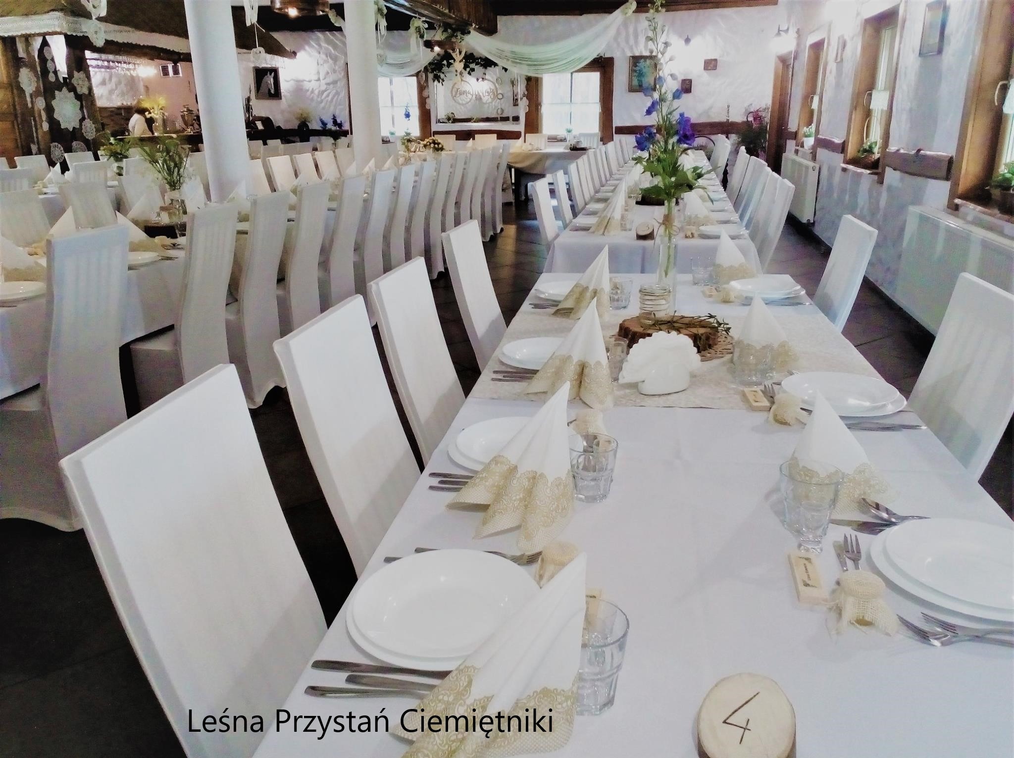 Uroczysta sala bankietowa z długimi stołami, nakryta białą zastawą i dekoracjami w stylu rustykalnym. Krzesła z białymi pokrowcami. Widoczne kwiaty i numeracja stołów.