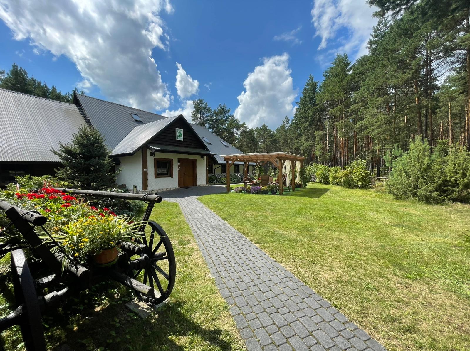 Dwór w leśnej scenerii z wózkiem kwiatów, ścieżką z kostki i drewnianą altaną. Idealne miejsce na imprezy okolicznościowe i catering.