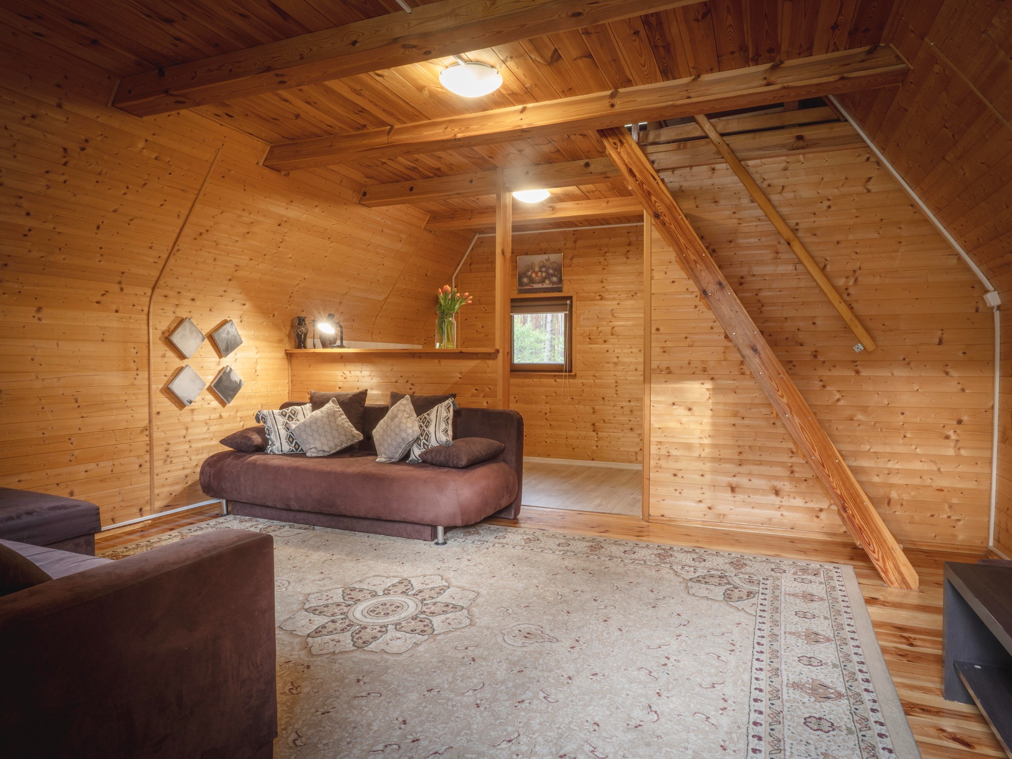 Drewniany salon z brązową sofą, wzorzystym dywanem i schodami. Ściany i sufit wyłożone boazerią. Na półce wazon z tulipanami. Przytulne wnętrze.