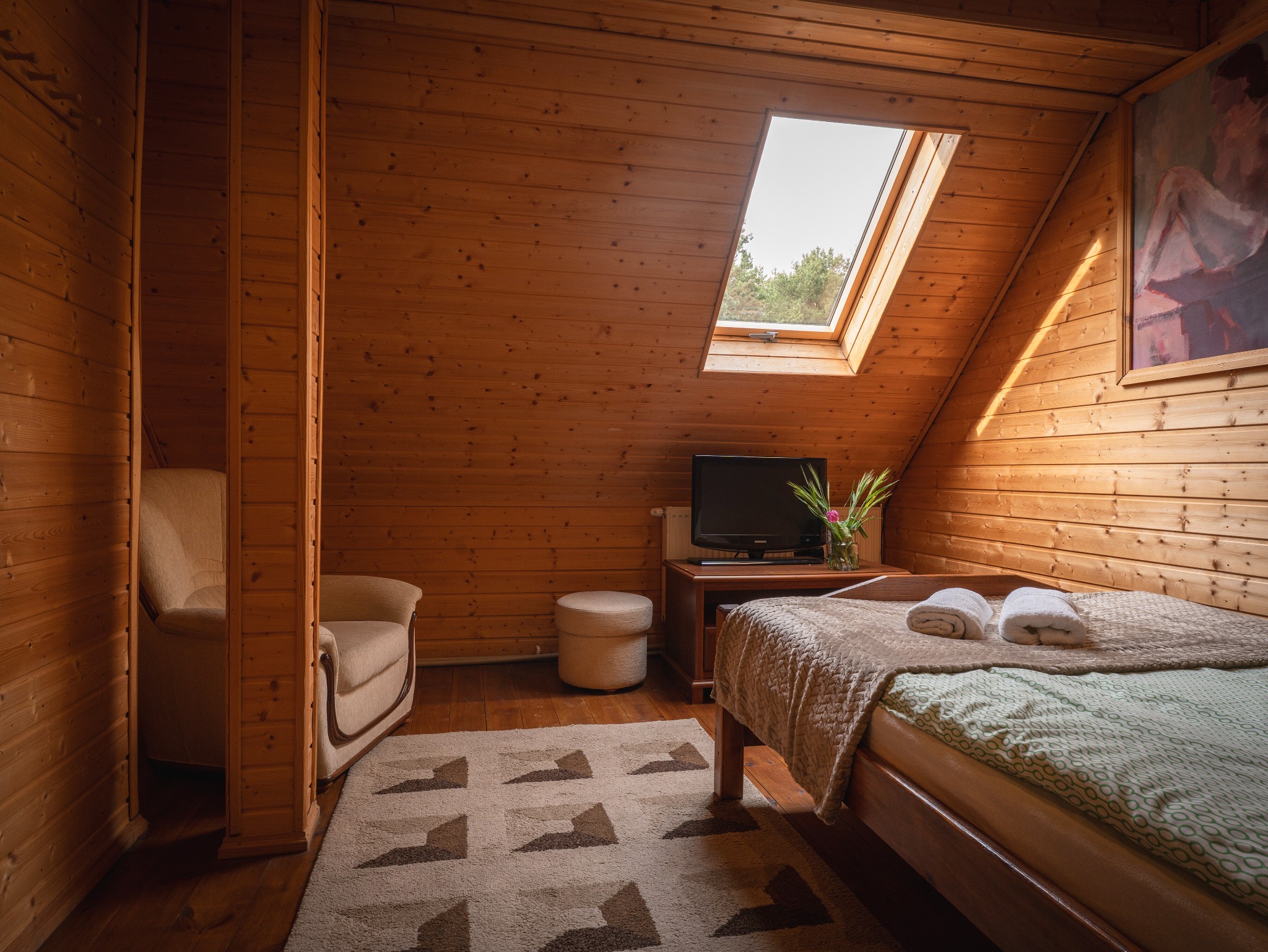 Przytulny pokój w drewnie z łóżkiem, telewizorem i oknem dachowym. Wazon z kwiatami dodaje uroku. Widoczny fotel i dywan. Całość tworzy ciepłą, domową atmosferę.
