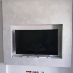 Wnęka na telewizor z dekoracyjną szarą ścianą, widoczne gniazdka elektryczne i fragment podłogi w trakcie montażu, z opakowaniem telewizora pod ekranem.