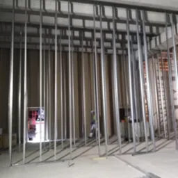 Montaż metalowego stelażu pod płytę karton gips na ścianie w surowym wnętrzu, widoczne profile podłogowe i sufitowe, osoba w tle.