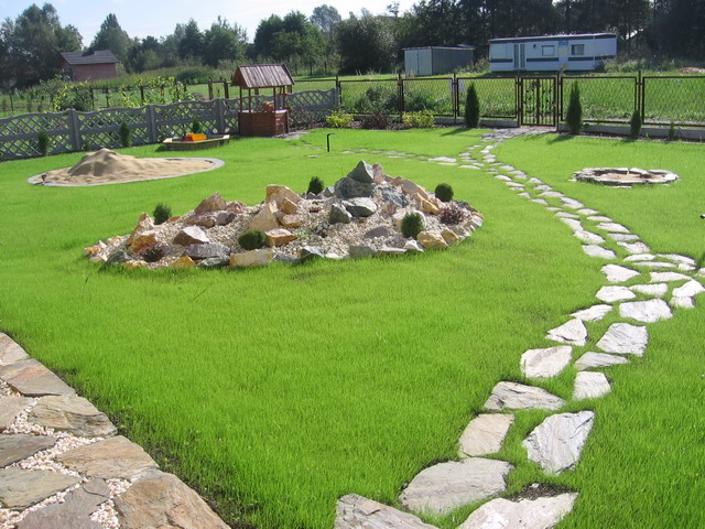 Zadbany ogród z trawnikiem, ścieżką z nieregularnych kamieni, skalniakiem z kamieni i żwiru, studnią, piaskownicą oraz ogrodzeniem w tle.