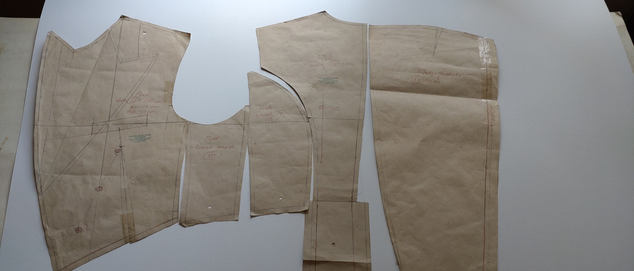 Rozłożone na białym stole papierowe wykroje fraka z naniesionymi odręcznymi notatkami i wymiarami, widoczne fragmenty wykroju przodu, boczka i tyłu.