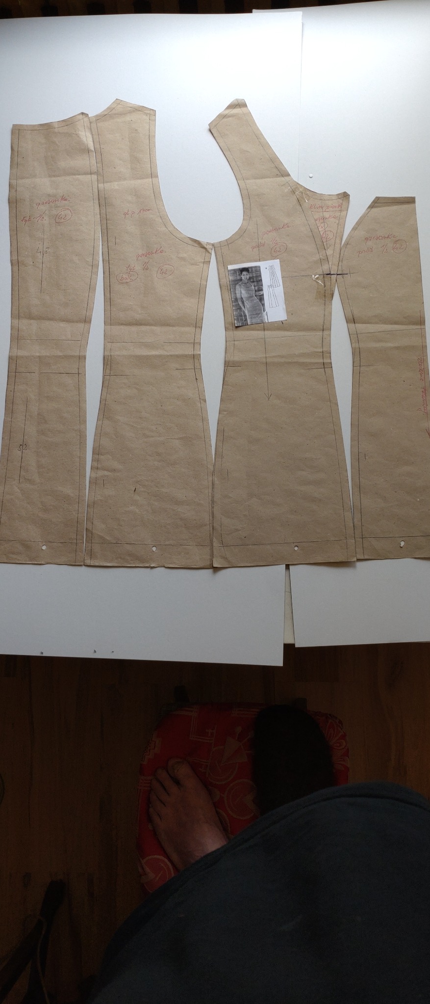 Papierowe wykroje sukienki damskiej z naniesionymi oznaczeniami, ułożone na białym stole, obok miniatura gotowej sukienki, widoczny fragment podłogi i stopy osoby siedzącej.