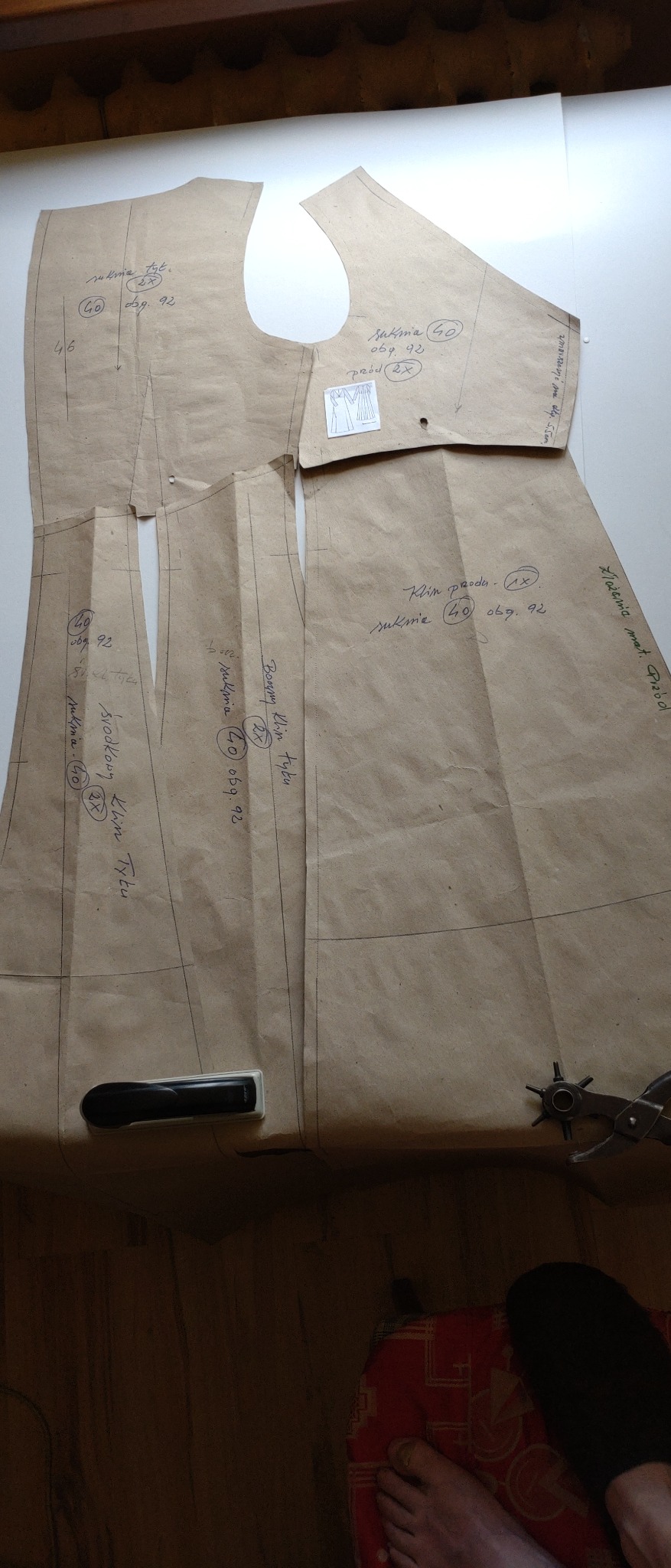 Papierowe wykroje sukienki z naniesionymi odręcznymi notatkami, leżące na stole obok dziurkacza krawieckiego i obciążnika, widok z góry.