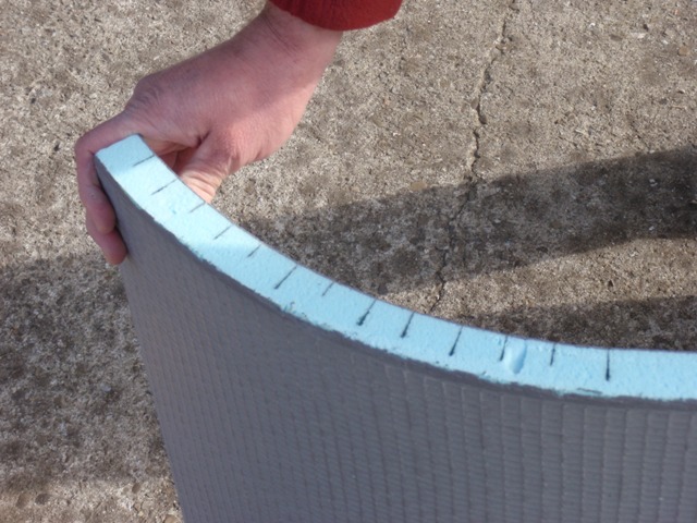 Ręka trzyma zakrzywiony fragment wodoodpornej płyty budowlanej z niebieskim rdzeniem i naniesionymi czarnymi oznaczeniami, na tle betonowej powierzchni.