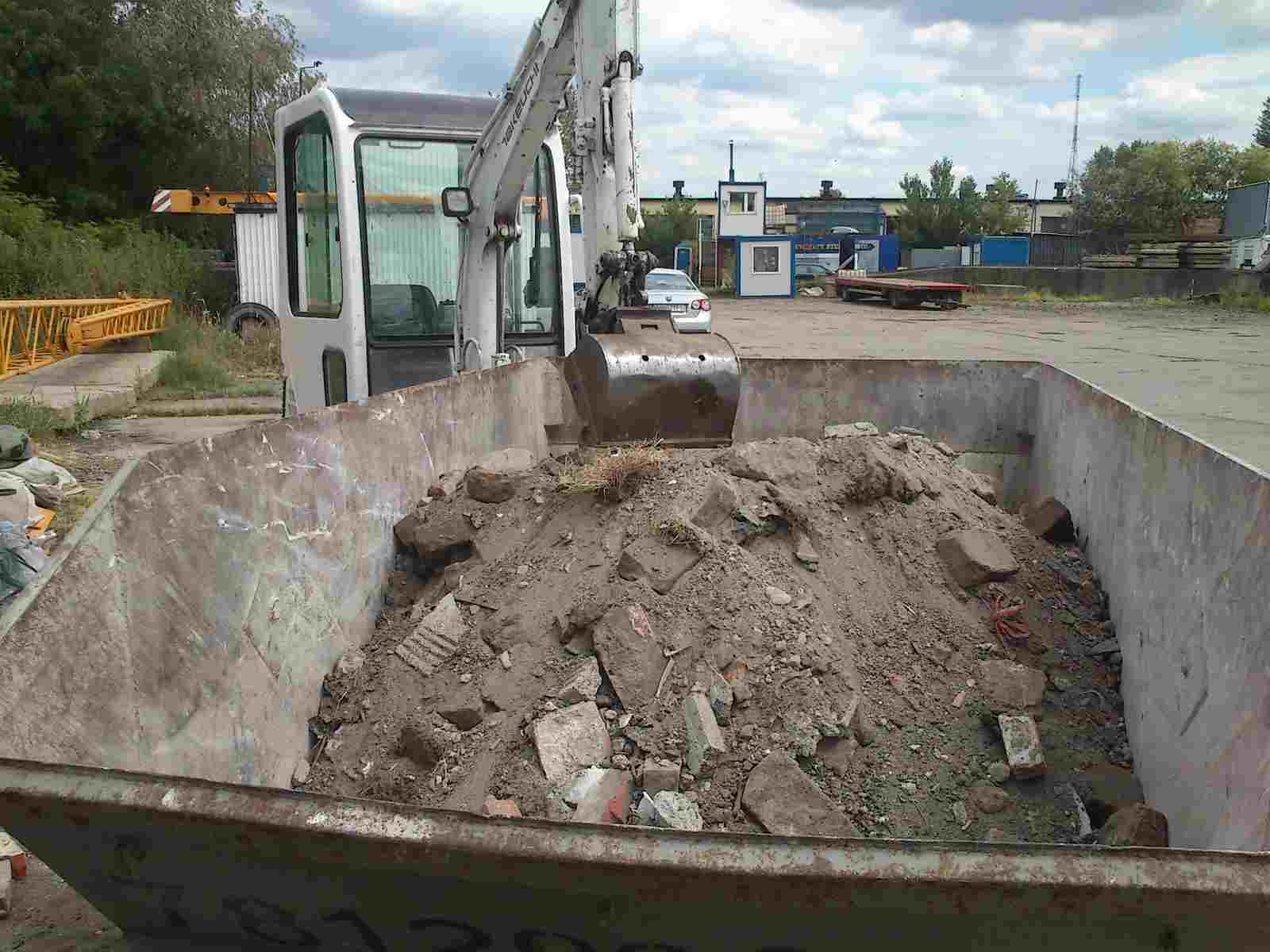 Mini koparka nabiera gruz i ziemię do kontenera, w tle budynek i zaparkowany samochód.