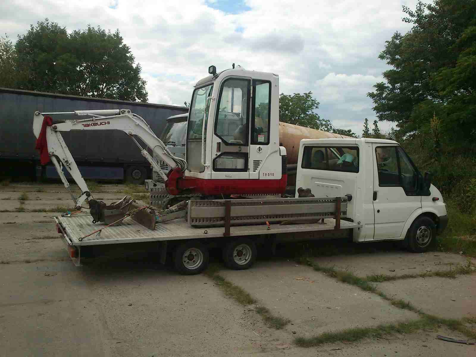 Biało-czerwona mini koparka Takeuchi TB016 na platformie transportowej białego Forda Transita, widok z boku na betonowym placu, w tle zaparkowane ciężarówki.