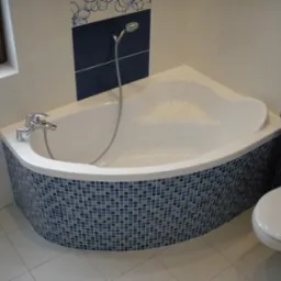 Biała wanna z mozaiką w odcieniach błękitu i granatu, dopasowana do płytek na ścianie. Nowoczesna armatura i kabina prysznicowa. Widoczna toaleta.