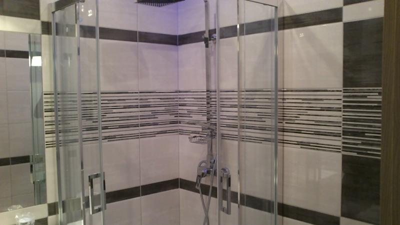 Nowoczesna kabina prysznicowa z chromowaną armaturą i szklanymi drzwiami na tle płytek w odcieniach bieli i szarości z dekoracyjnymi pasami.