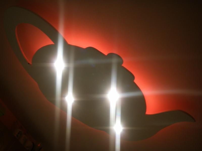 Dekoracyjna lampa ścienna w kształcie czajnika, podświetlona na czerwono, z wyraźnymi refleksami świetlnymi.