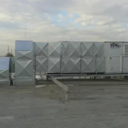 Instalacja wentylacyjna VBW na dachu budynku, widok z boku. Metalowe elementy o kwadratowej strukturze, instalacja na platformie. Pochmurne niebo w tle.