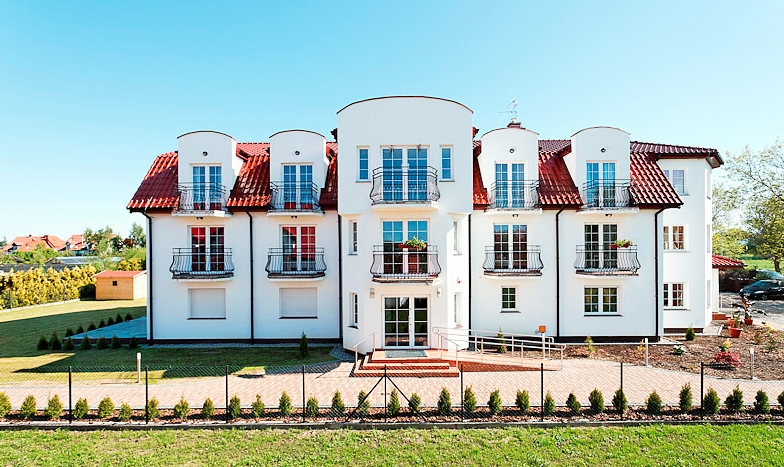 Nowoczesny, trzypiętrowy budynek z białymi ścianami, czerwonym dachem i balkonami z czarnymi balustradami, otoczony zielonym trawnikiem i niskim ogrodzeniem z tujami.