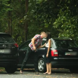 Para całująca się między dwoma zaparkowanymi samochodami na tle gęstej zieleni.