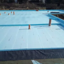 Izolacja termiczna z płyt styropianowych na fundamencie, widoczne pionowe rury instalacyjne i czarna folia hydroizolacyjna.