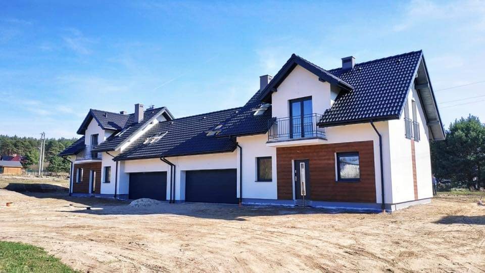Nowoczesne segmenty bliźniacze w stanie deweloperskim, z czarną dachówką, białymi ścianami i drewnianą elewacją, widoczne garaże i balkon z czarną balustradą.