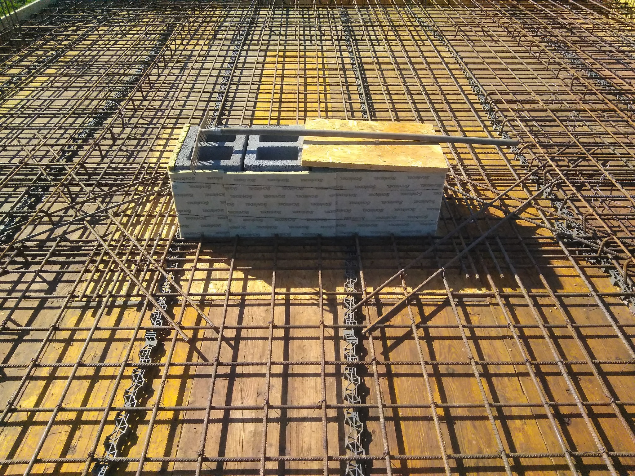 Zbrojenie fundamentu z siatką stalową na żółtej płycie szalunkowej, z ułożonymi bloczkami betonowymi i deską, przygotowane do zalania betonem.