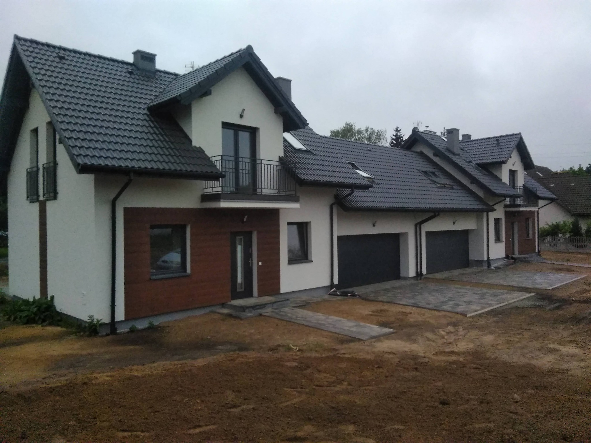 Szeregówka domów murowanych z ciemną dachówką i garażami, widok z zewnątrz na etapie zagospodarowania terenu.