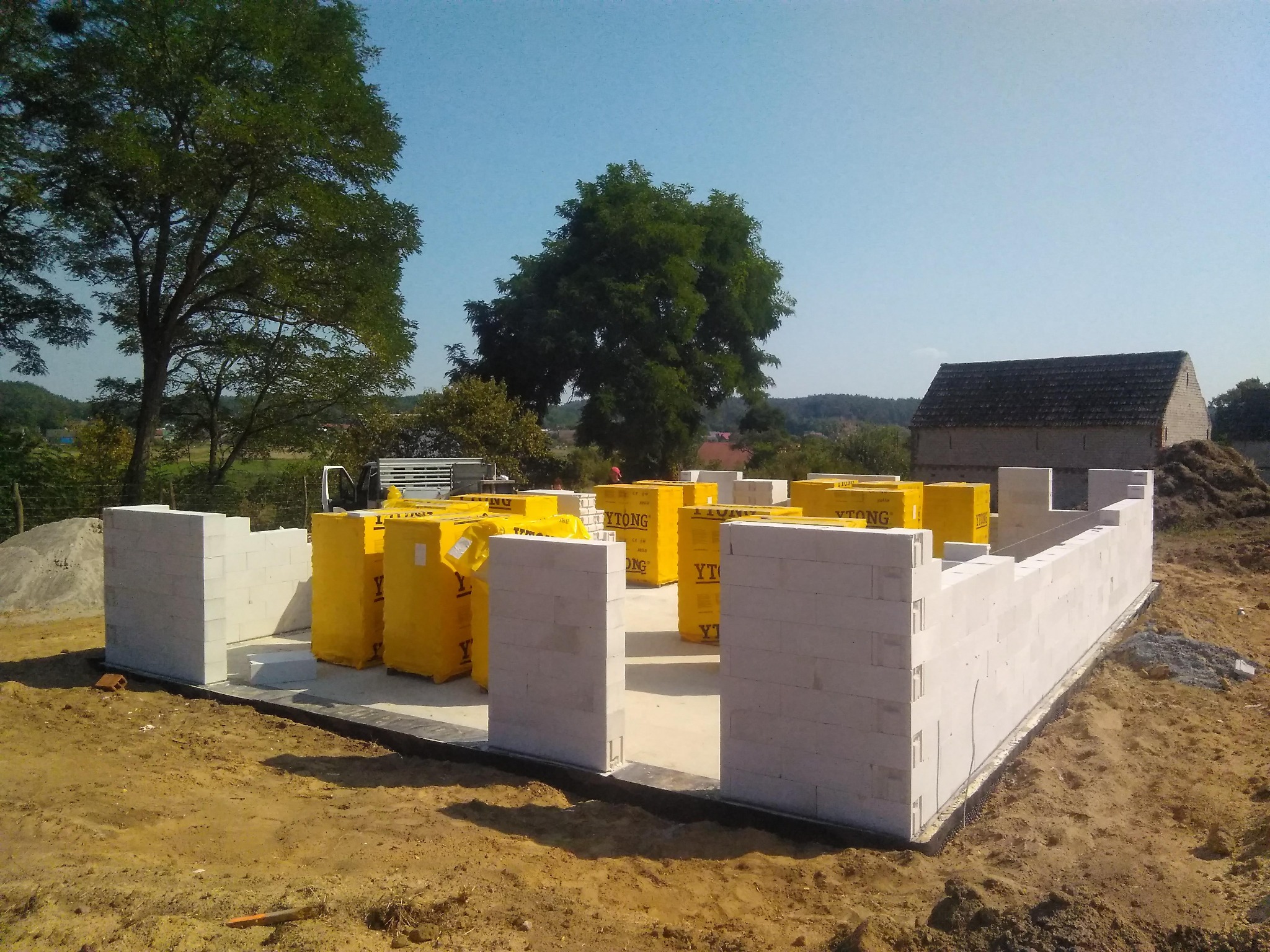 Wznoszenie ścian parteru z bloczków Ytong na przygotowanym fundamencie. Na paletach widać zapakowane bloczki. W tle drzewa i budynek gospodarczy.