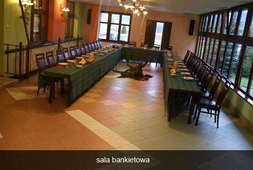 Ustawiona sala bankietowa z długim stołem w kształcie litery U, nakrytym zielonym obrusem, z zastawą i dekoracjami, w tle okna i ściany w ciepłych kolorach, widoczne krzesła i oświetlenie.