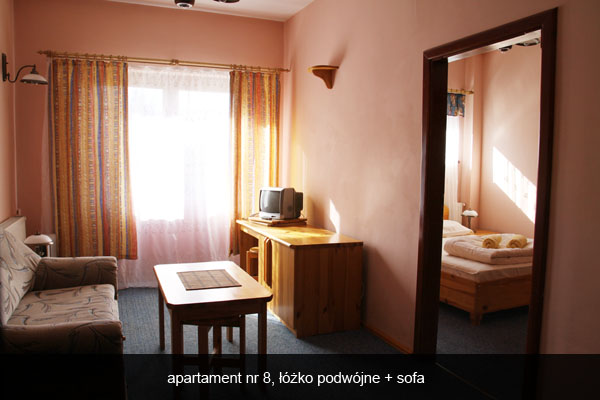 Pokój hotelowy z sofą, stolikiem kawowym i biurkiem z telewizorem, widok na sypialnię z podwójnym łóżkiem przez otwarte drzwi.