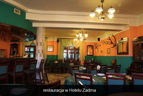 Wnętrze restauracji hotelowej z barem, stolikami nakrytymi zielonymi obrusami, dekoracjami z poroża na pomarańczowych ścianach i oświetleniem w stylu retro.