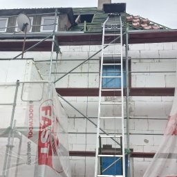 MP-BUD Usługi Budowlane - Pracownik w kasku stoi u podnóża drabiny opierającej się o budynek w trakcie prac remontowych dachu, widoczne rusztowania i materiały izolacyjne z logo firmy FAST.
