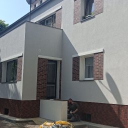 MP-BUD Usługi Budowlane - Pracownik maluje dolną część elewacji budynku, widoczne narzędzia malarskie, pędzle, wiaderka z farbą i skrzynka narzędziowa, budynek z oknami w ceglanej oprawie i anteną satelitarną na dachu.