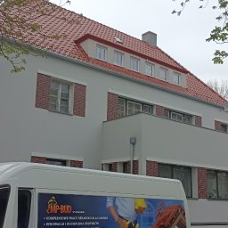 MP-BUD Usługi Budowlane - Odnowiony budynek z nową czerwoną dachówką, białą elewacją i ceglanymi obramowaniami okien, częściowo zasłonięty przez drzewa i pojazd firmy budowlanej z reklamą usług dekarskich i dociepleń.
