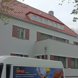 Odnowiony budynek z nową czerwoną dachówką, białą elewacją i ceglanymi obramowaniami okien, częściowo zasłonięty przez drzewa i pojazd firmy budowlanej z reklamą usług dekarskich i dociepleń.