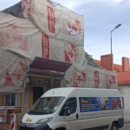 MP-BUD Usługi Budowlane - Budynek w trakcie prac dekarskich, zabezpieczony folią z logo producenta materiałów izolacyjnych, widoczne krokwie dachu, palety z dachówkami na pierwszym planie i samochód dostawczy firmy dekarskiej.