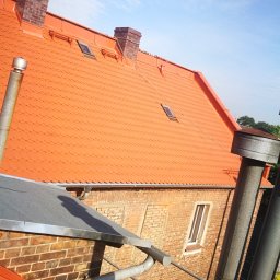 MP-BUD Usługi Budowlane - Czerwony dach z dachówki karpiówki na ceglanym budynku, widoczne kominy z cegły klinkierowej, okna dachowe, rynna i fragment rusztowania na pierwszym planie, błękitne niebo w tle.