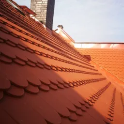 Nowy dach pokryty dachówką karpiówką w odcieniach czerwieni, z widocznym kominem z cegły i fragmentem nieba z lekkimi chmurami.