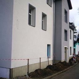 MP-BUD Usługi Budowlane - Dwukondygnacyjny budynek po ociepleniu elewacji, widoczne świeżo otynkowane ściany w odcieniach szarości i białe obramowania okien, teren wokół budynku częściowo rozkopany, zabezpieczony taśmą.