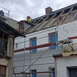 MP-BUD Usługi Budowlane - Budynek w trakcie prac remontowych: ocieplanie elewacji styropianem, częściowo zdjęte pokrycie dachowe, widoczne rusztowania, pracownicy na dachu, okna zabezpieczone folią.