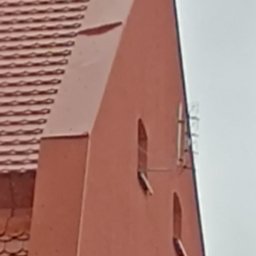 MP-BUD Usługi Budowlane - Czerwony budynek z dwoma wąskimi oknami i anteną na ścianie, widoczny fragment dachu z dachówką karpiówką.